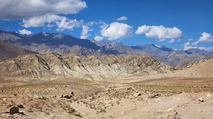 Layered desert ridges with sweeping striations under a bright blue sky dotted with cumulus clouds.