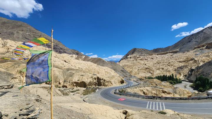 A ribboning highway curves through yellow lunar-like badlands with a fluttering prayer flag in the foreground.