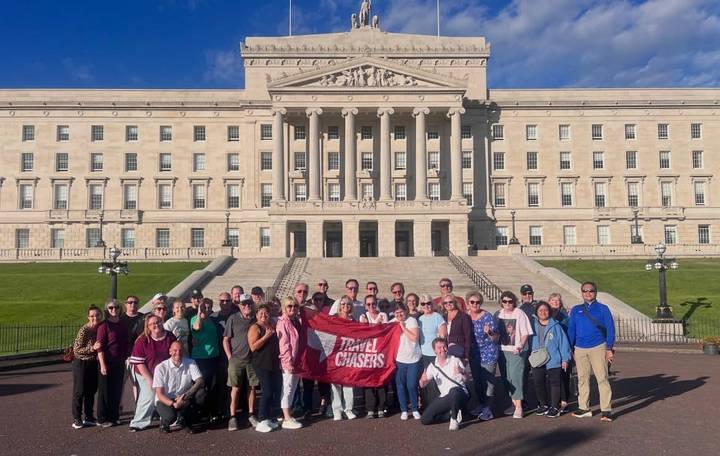 Large tour group posing with a red banner on the steps of a neoclassical government building under blue skies