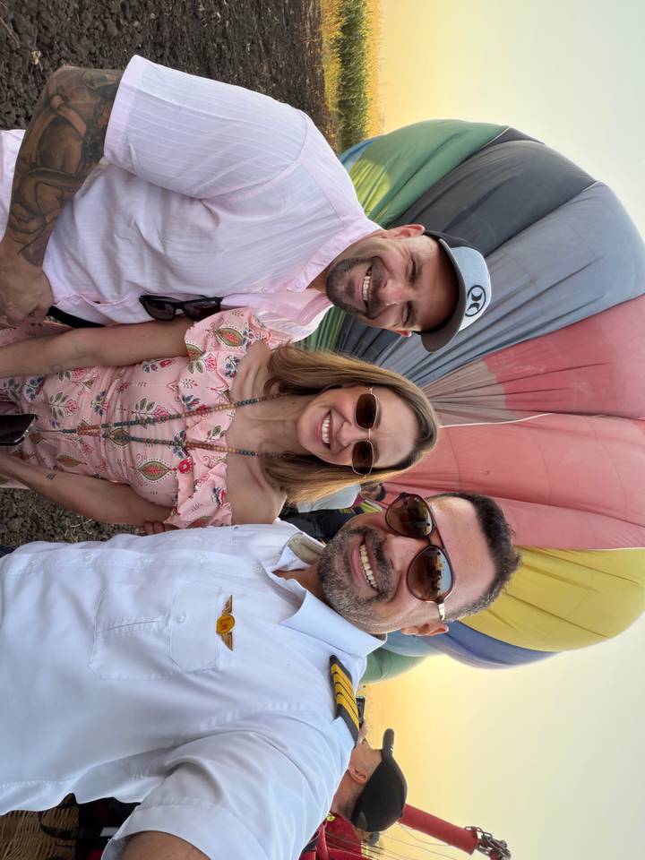 Three friends smiling in front of a colorful inflated hot-air balloon envelope.