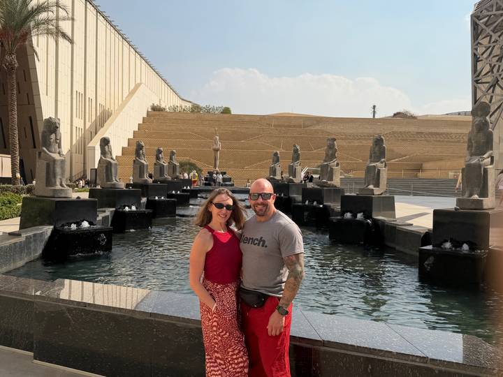 Couple posing by modern fountains lined with pharaonic statues outside a grand museum entrance.