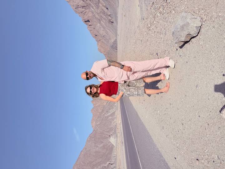 Travelers stand barefoot on a lonely desert highway flanked by rugged brown mountains under clear blue skies.