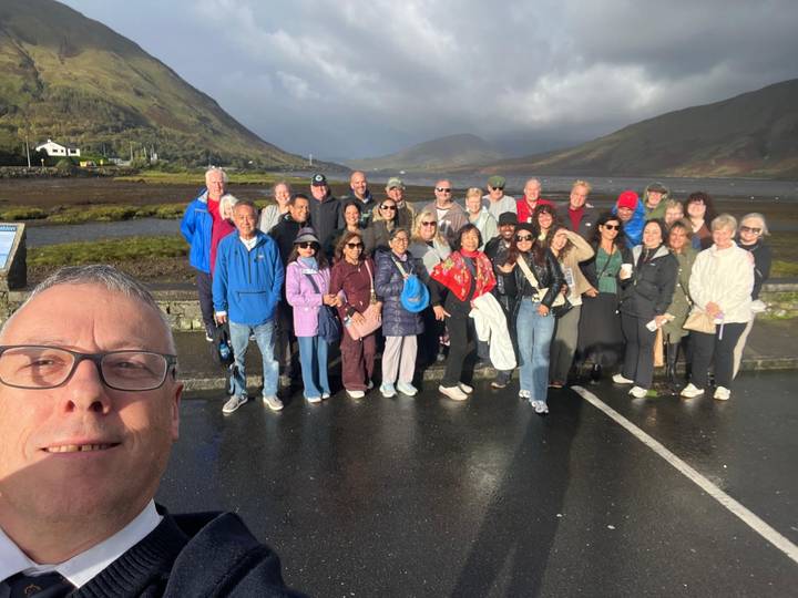 Large tour group taking a cheerful selfie in front of misty Irish mountains and a lake.