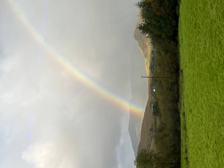 Vivid rainbow arcs over green Irish countryside with distant mountains under stormy clouds.