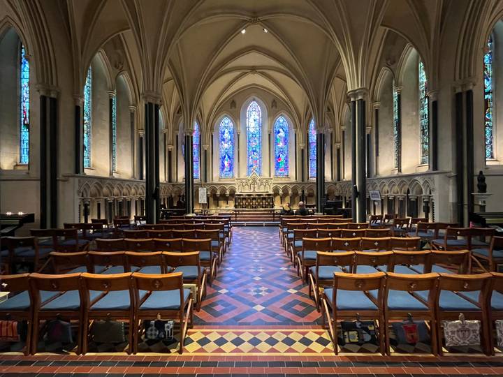 Elegant Gothic cathedral nave with patterned tile floor, wooden chairs, and colorful stained glass windows.