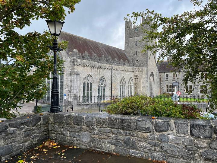 Stone church with tall arched windows seen behind leafy branches and old stone wall.