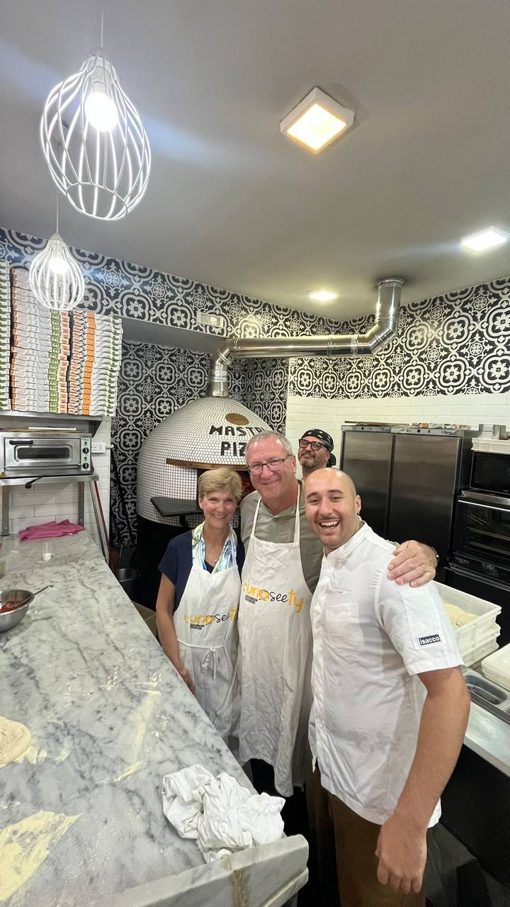 Cooking-class participants pose with a chef inside a pizzeria featuring a tiled pizza oven.