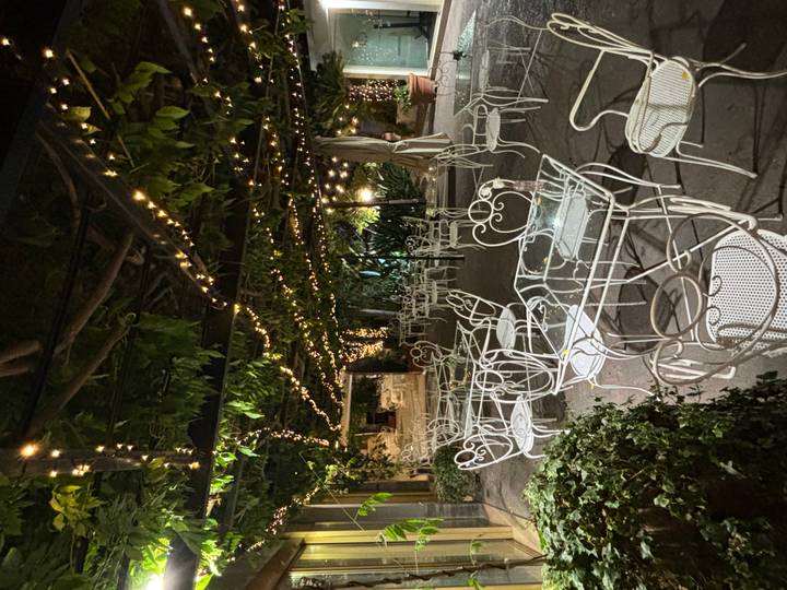 Charming outdoor café with white wrought-iron furniture and fairy lights strung beneath leafy vines at night.