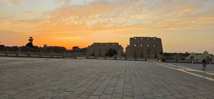 Orange sunset sky behind the vast courtyard and ruins of Karnak Temple viewed across a paved plaza.