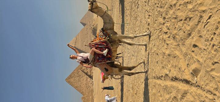Traveler riding a decorated camel in the desert with the Pyramids of Giza rising in the background.