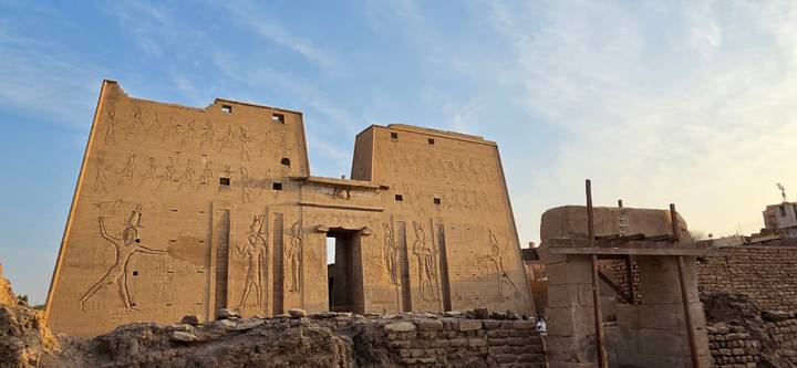 Sandstone walls of Edfu Temple covered in reliefs, lit by warm late-day sunlight with some restoration scaffolding nearby.