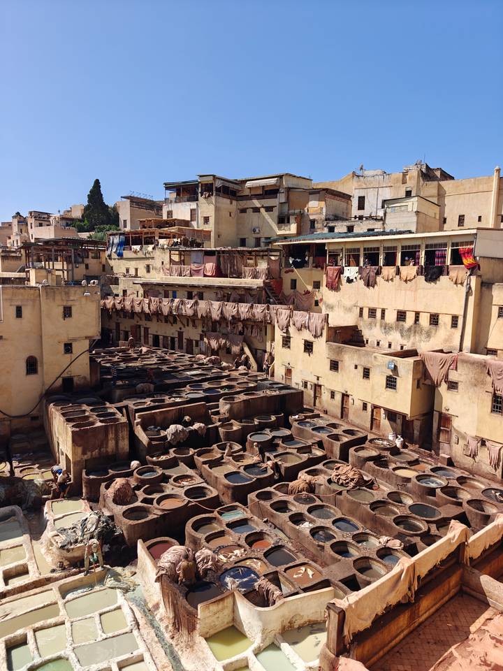 View over the Chouara Tannery in Fes with numerous stone dye vats, hanging leather skins and surrounding tan buildings under bright sunlight.