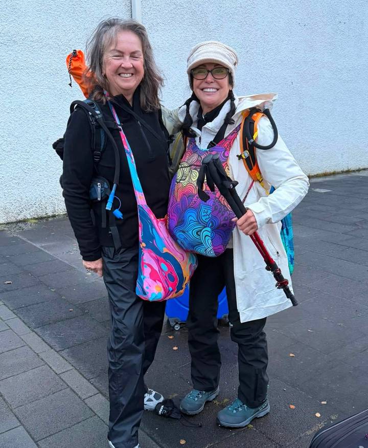 Two equipped hikers smile on a city pavement with backpacks and colourful patterned bags.