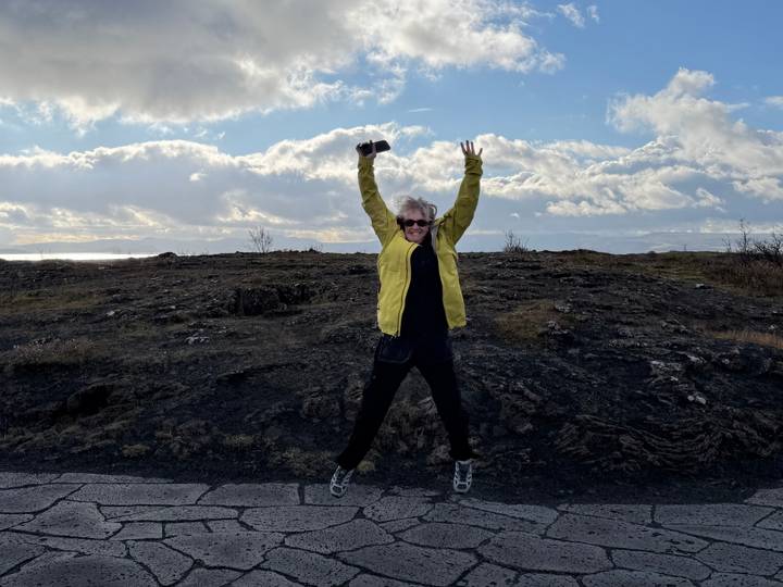 Person in yellow jacket jumps with arms raised on a barren lava plain under dramatic clouds.