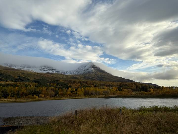 Autumn forest colours blanket lowlands beneath a mist-capped pyramid mountain beside a still lake.