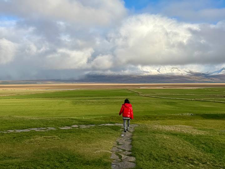 Lone hiker in a red jacket walks a stone path across vast green plains towards snowy mountains.