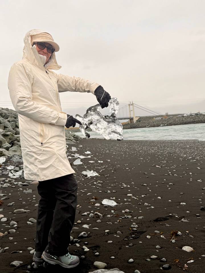 Traveller in light coat holds a clear shard of glacial ice on a black-sand beach.