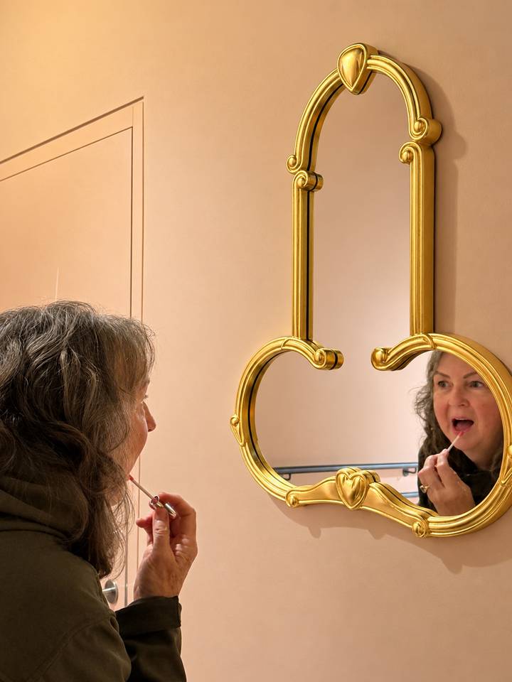 Woman applies lipstick while looking into a decorative gold mirror in an indoor setting.