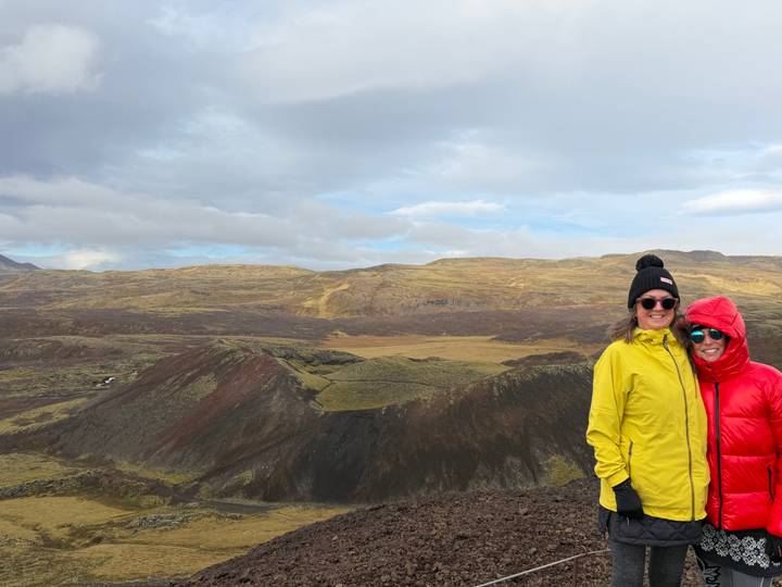 Two friends in colourful jackets stand on the rim of a volcanic crater under a cloudy sky.