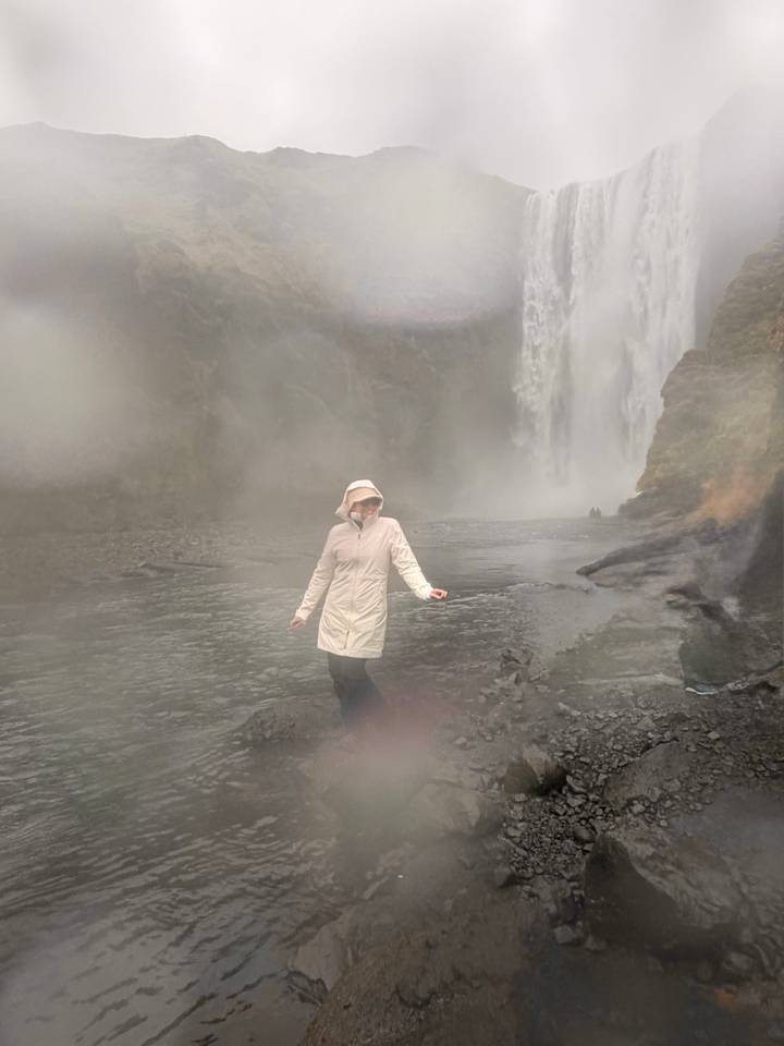 Traveller in raincoat stands in heavy mist before a tall waterfall, surroundings blurred with spray.