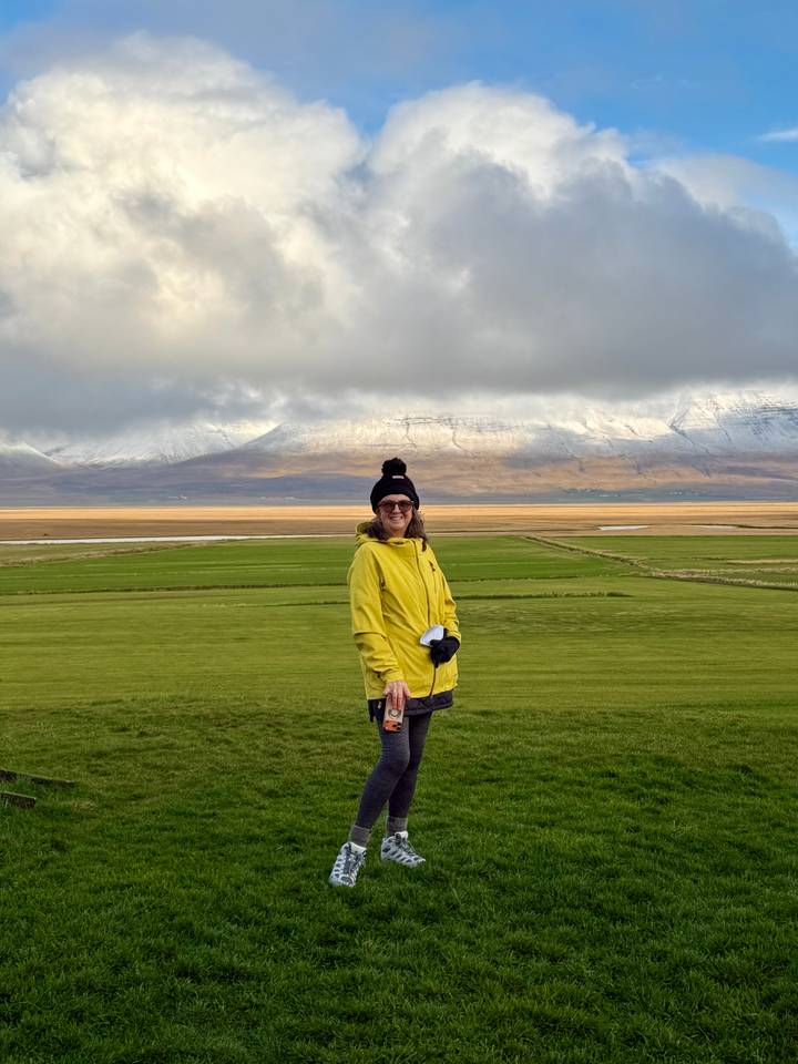 Smiling traveller in yellow jacket stands on rolling green fields with snow-capped peaks behind.