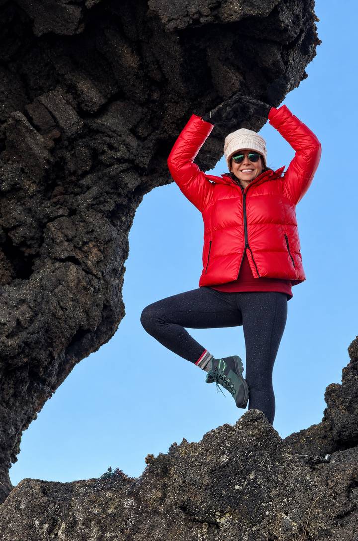 Close-up of climber hanging playfully from a dark lava arch against a clear sky.