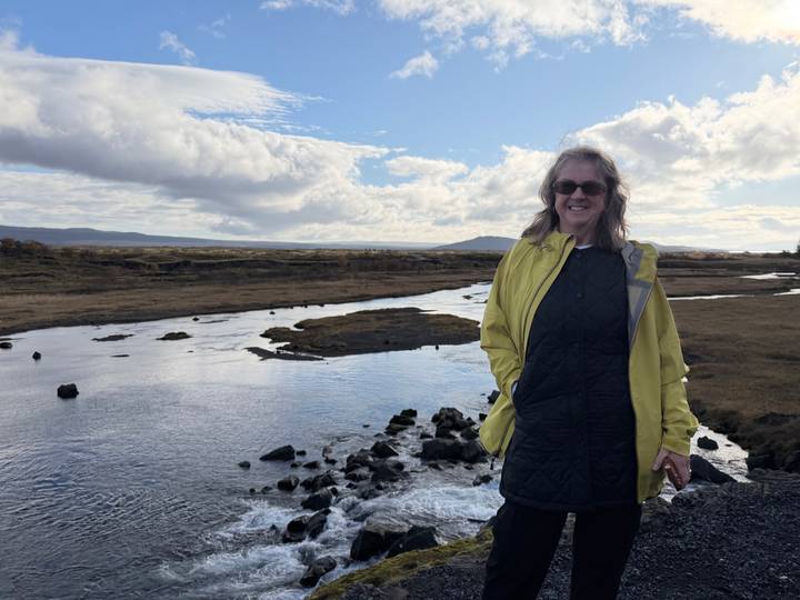 Traveller in yellow jacket stands beside a reflective river under a patchy cloud sky.