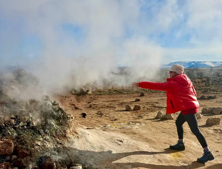 Red-jacketed visitor extends arms toward steaming fumarole on a sun-lit geothermal plain.