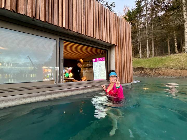 Guest enjoys a swim-up bar in a forest lagoon while staff serves drinks through a window.