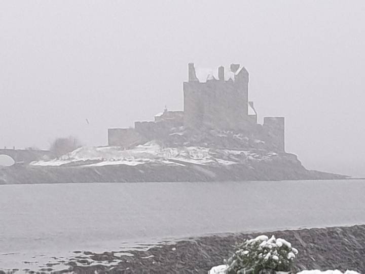 Silhouetted castle on an islet barely visible through heavy snowfall and grey mist across a cold loch.