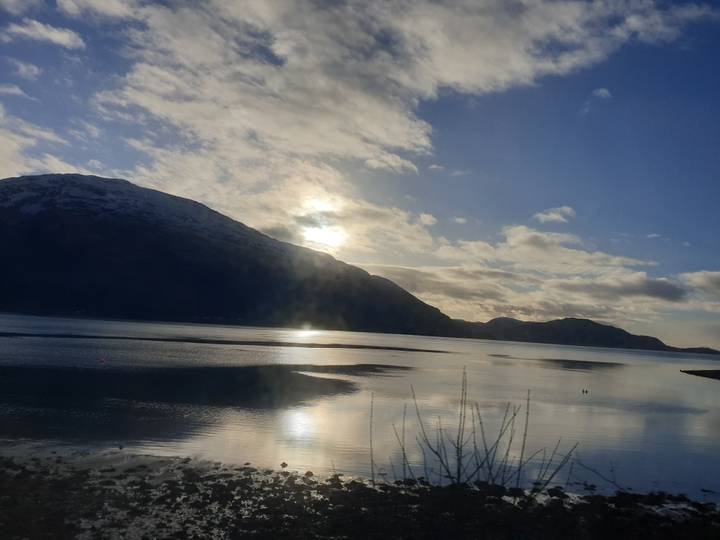 Serene Scottish loch reflecting low winter sun with dark mountains and scattered clouds on the horizon.