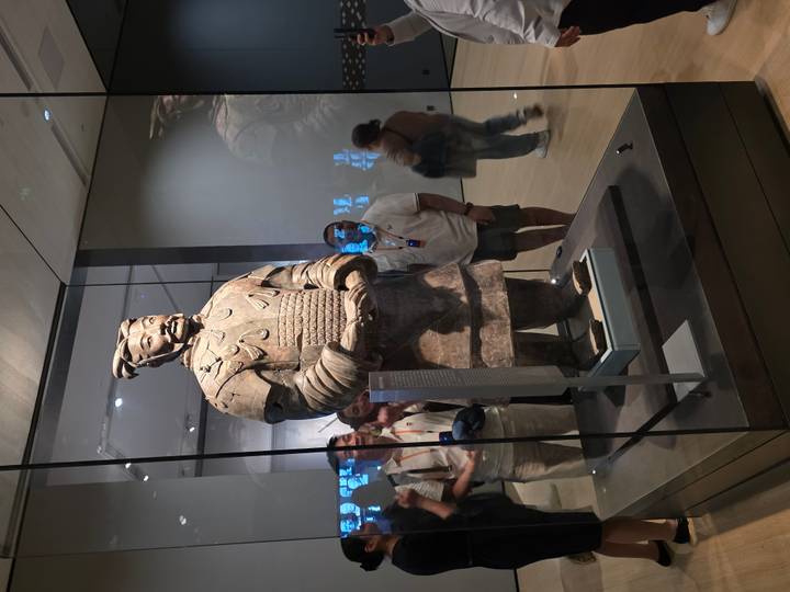 Terracotta Warrior statue displayed in a glass case surrounded by museum visitors taking photos.
