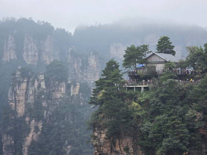 Cliff-edge teahouse perched among sheer sandstone spires and lush greenery under a misty sky in Zhangjiajie.