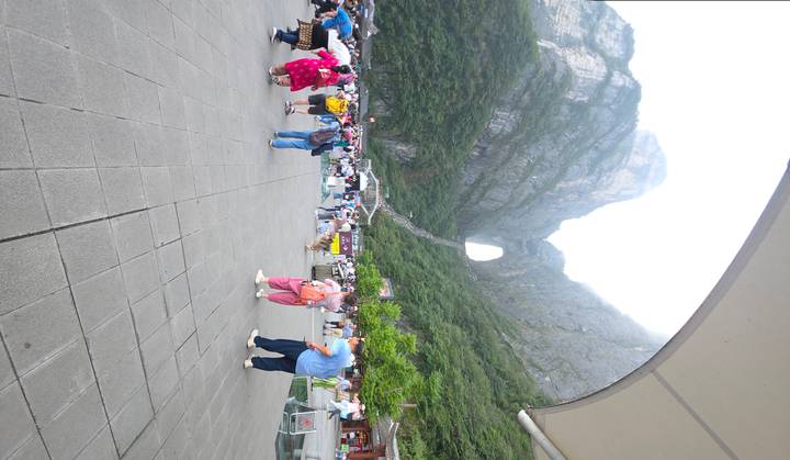 Wide plaza full of tourists looking toward the giant natural arch of Tianmen Mountain rising steeply behind a long staircase.
