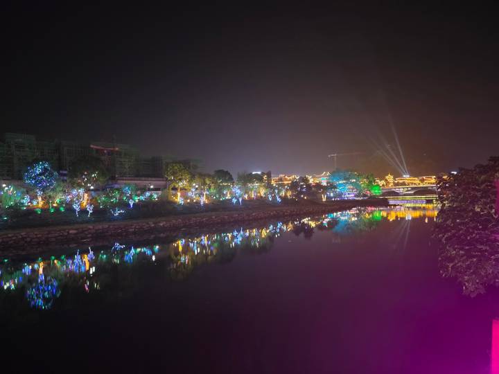 Colorful riverside light display reflecting in calm water against a night sky with searchlights beaming upward.
