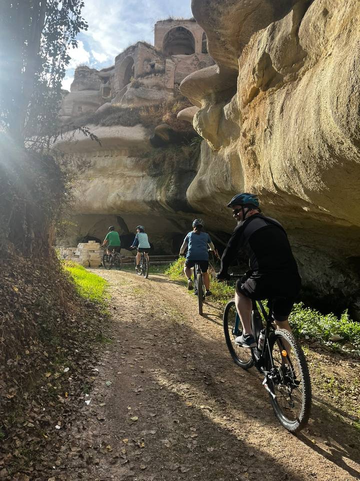 Cyclists ride single file toward a cave entrance carved in soft Cappadocian rock, sunlight streaming through trees.