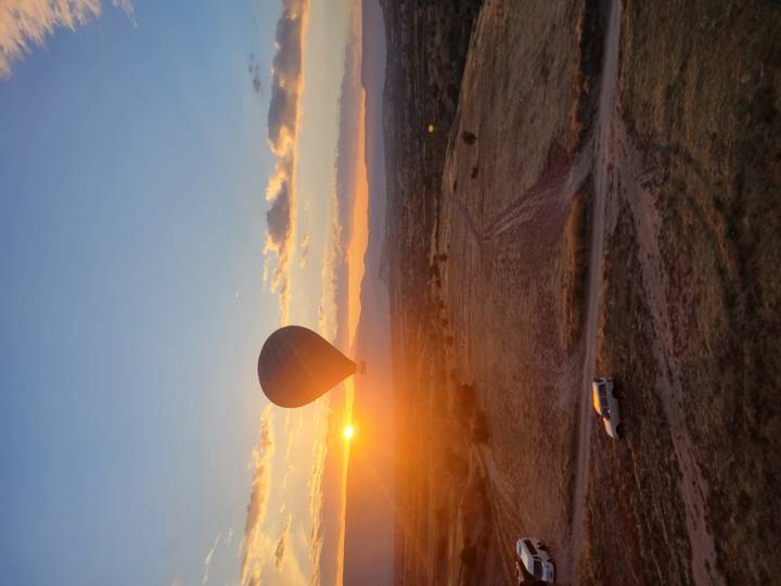Silhouette of a hot-air balloon floating above Cappadocia at sunrise with golden sun on the horizon and a lone vehicle on a dirt track below.