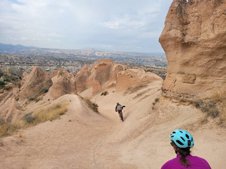Mountain biker descends a sandy ridge between rose-colored rock fins while another cyclist looks on.