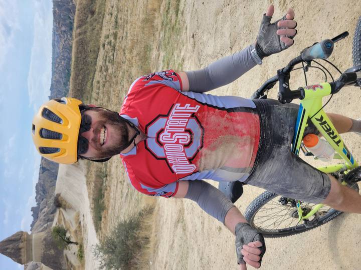 Dust-covered cyclist in Ohio State jersey smiles holding his mountain bike after a ride on Cappadocian trails.