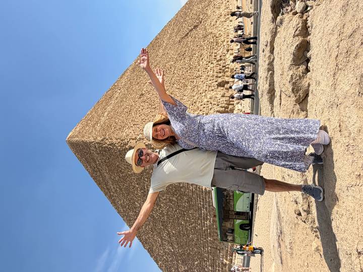 Smiling couple with arms outstretched stand before the Great Pyramid of Giza on a clear blue day.