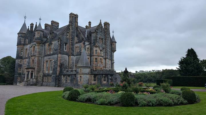 A stately stone manor with turrets overlooks manicured gardens under a gray sky.