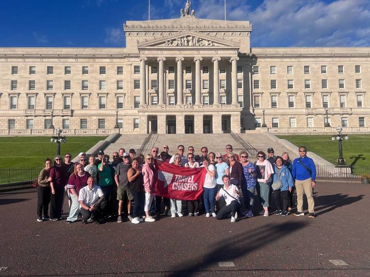 A large tour group poses with a Travel Chasers banner on the steps of Stormont Parliament in Belfast.