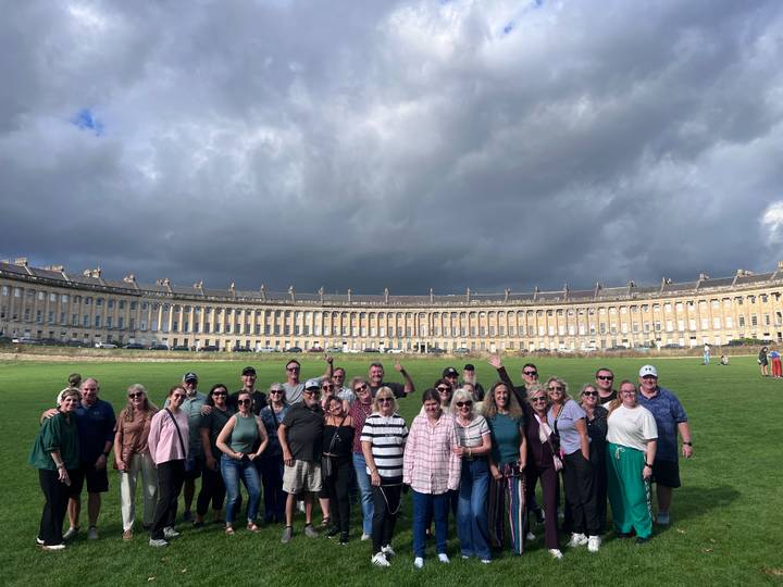 A tour group smiles before the sweeping Georgian arc of Bath’s Royal Crescent under dramatic skies.