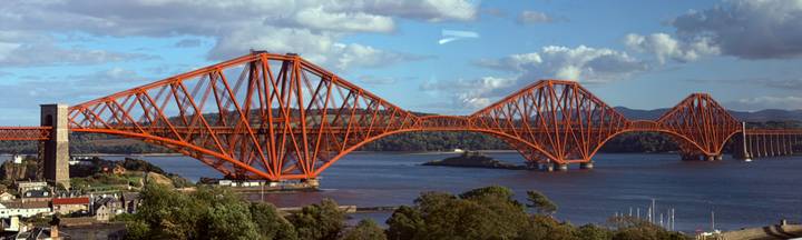 Bright red cantilever railway bridge stretching across a wide estuary under a blue sky with scattered clouds.