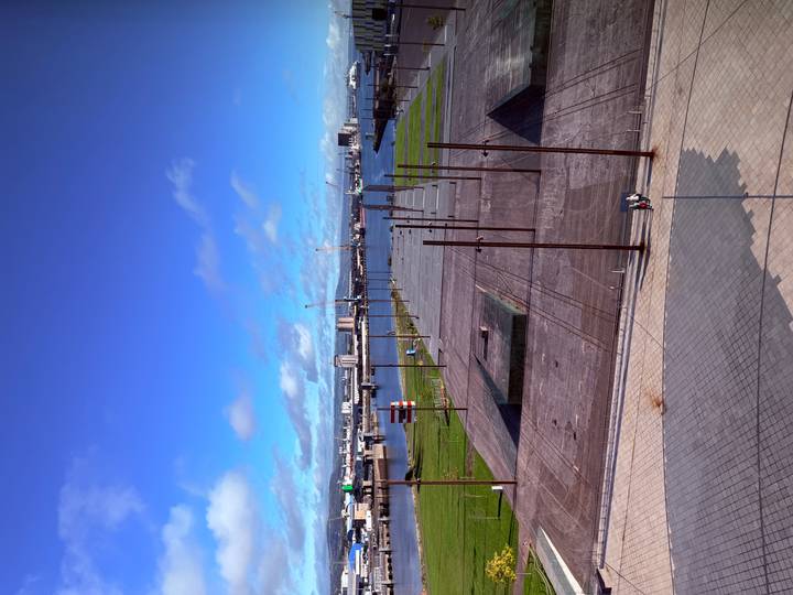 Wide open urban waterfront plaza with rows of metal poles facing a harbor dotted with cranes and distant buildings.