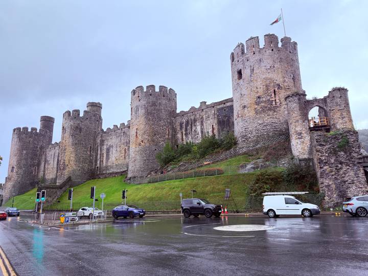 Massive medieval stone castle with circular towers rising above a rainy street and lush green embankment.