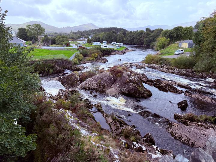 Rocky river rapids winding through lush countryside with scattered campers and rolling hills in the distance.