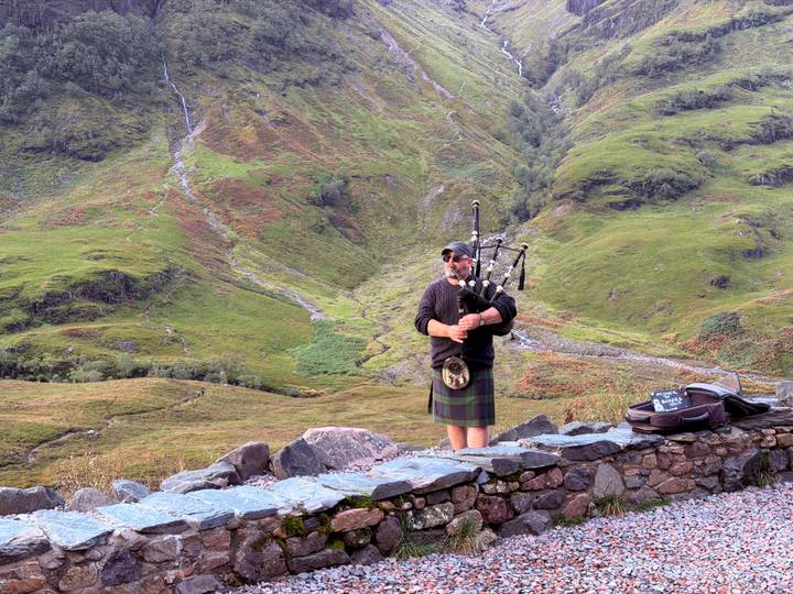 Traditional Scottish bagpiper in kilt performing on a stone wall with verdant Highlands backdrop.