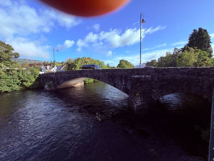 Stone arch bridge carrying a narrow road over a dark, flowing river surrounded by leafy trees.