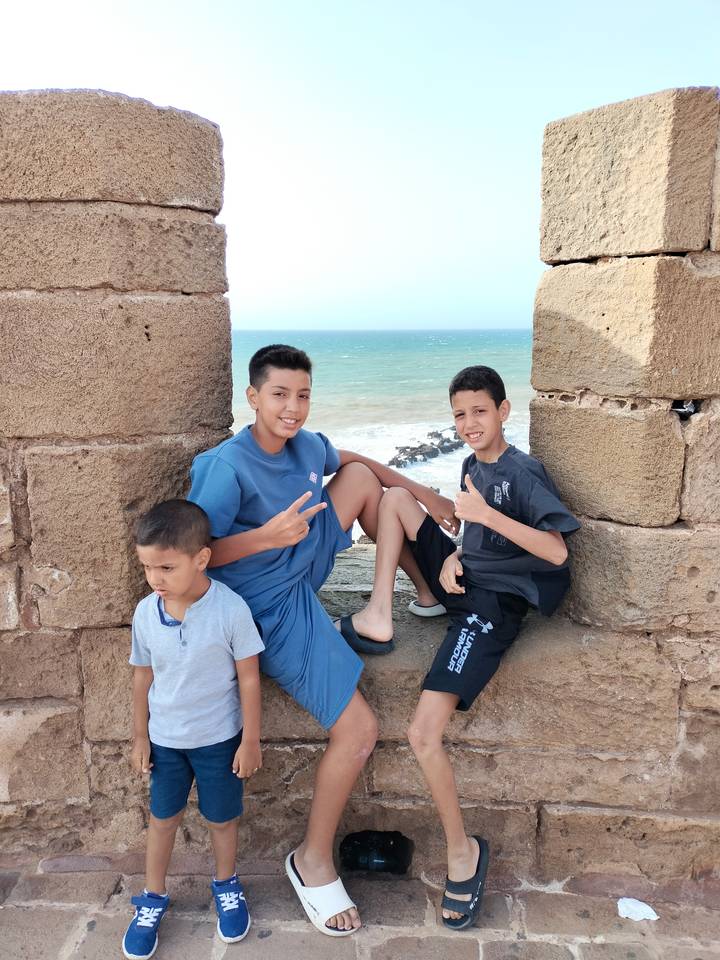 Three boys sit and smile in a stone window opening overlooking the turquoise Atlantic Ocean.